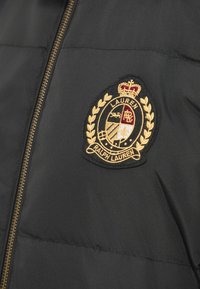 Melna spīdīga jaka ar rāvējslēdzēja aizdari un zeltītu izšūtu apļveida logo ar lauru lapu uzlikām un kroni kreisajā krūtīs.