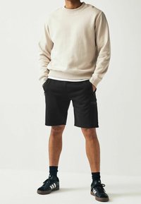 Sweat-shirt beige avec col rond côtelé, associé à un short cargo noir avec poches zippées latérales, et des baskets noires avec des accents blancs.