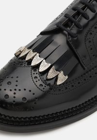 Scarpe da uomo di pelle nera con accenti decorativi in argento, dettagli brogue perforati e strisce di pelle frangiata sopra i lacci.
