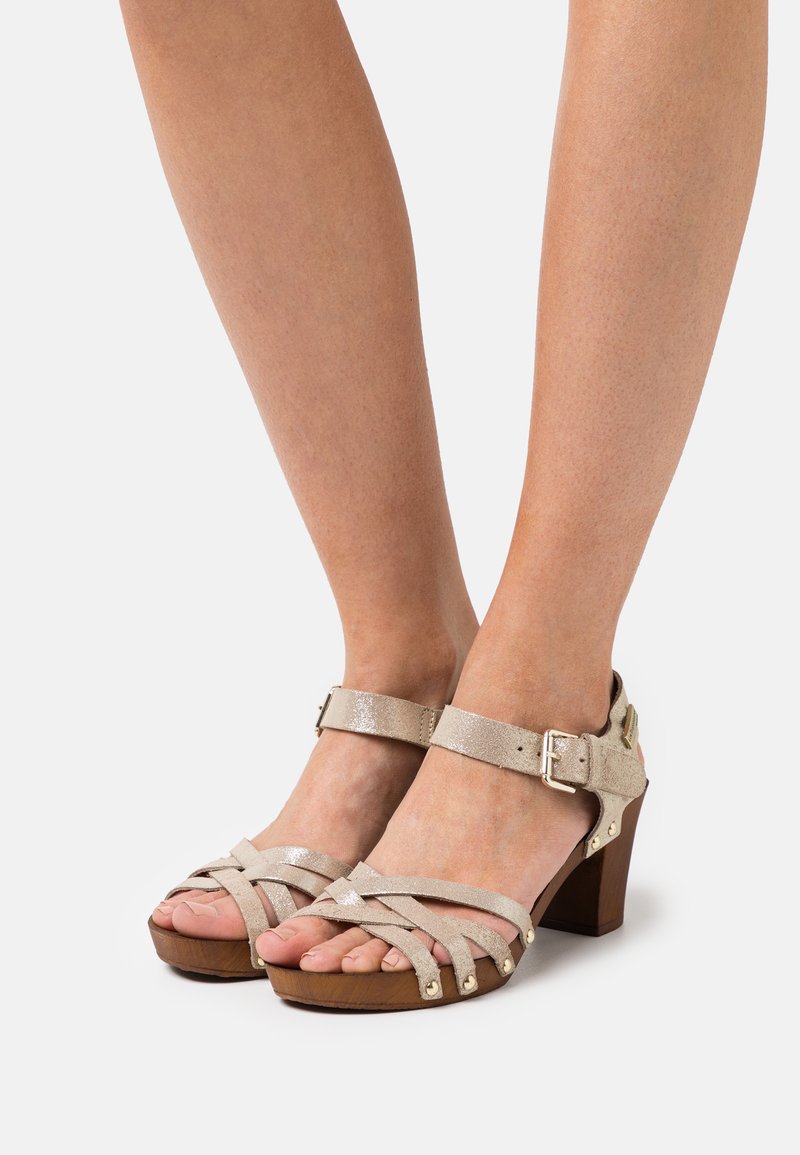 Beige strappy sandals with a textured finish, ankle buckle, and wooden heel. Features crossed straps and metallic accents on the sole.