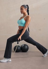 Athletic outfit featuring a light blue sports bra and black flared leggings. The individual holds a resistance band, with kettlebells in the background.