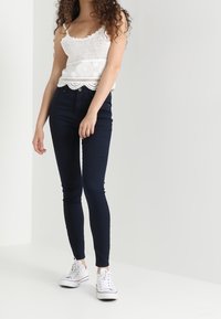 Vrouw met krullend haar, gekleed in een witte gehaakte tanktop, donkere spijkerbroek en witte sneakers, staand tegen een effen lichte achtergrond.