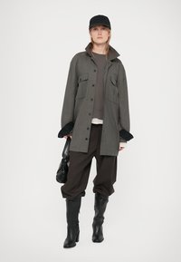 Γκρι πουκάμισο σακάκι με δύο μπροστινές τσέπες, τοποθετημένο πάνω από ένα σκούρο τοπ και κρέμα μπλούζα, συνδυασμένο με φαρδιά σκούρα παντελόνια και μαύρες μπότες μέχρι το γόνατο.