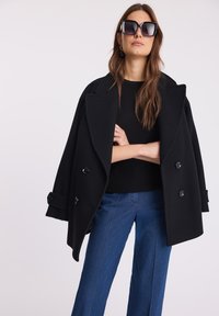 Czarny dwurzędowy wełniany płaszcz z dużymi klapami, noszony na czarnym swetrze i zestawiony z niebieskimi dżinsami o szerokich nogawkach. Czarne okulary przeciwsłoneczne dopełniają stylizację.