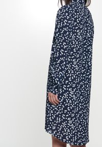 Abito navale con pois bianchi, maniche lunghe, colletto, polsini con bottone e vestibilità ampia. Presenta un dettaglio della cucitura posteriore e un orlo leggermente curvo.