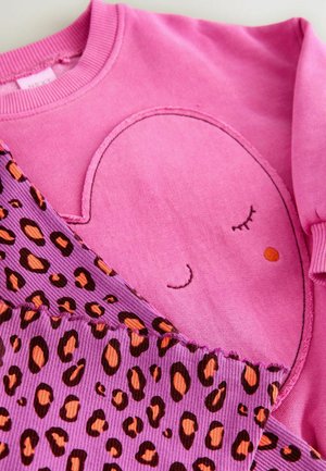 Sweatshirt rose avec un design de visage souriant au centre, associé à un tissu en velours côtelé violet présentant un imprimé léopard orange et marron.
