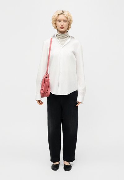 Άτομο με κοντά ξανθά μαλλιά που φοράει λευκό πουκάμισο πάνω από ριγέ ζιβάγκο, μαύρο παντελόνι, μαύρα παπούτσια και κρατάει ροζ τσάντα ώμου.