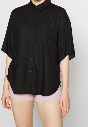 Kobieta ubrana w luźną czarną koszulę na guziki z krótkim rękawem i kieszenią na piersi, w połączeniu z jasnoróżowymi szortami z postrzępionymi krawędziami.