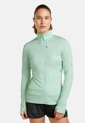 Woman wearing a mint green zip-up jacket with thumb holes and black athletic shorts, standing against a plain white background.