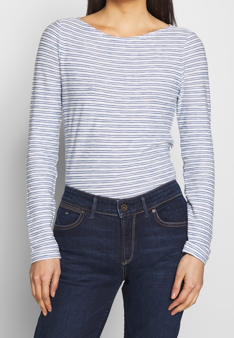 Frau in einem eng anliegenden weißen Langarmhemd mit dünnen blauen horizontalen Streifen und dunkelblauen Jeans von der Taille bis zur Mitte des Oberschenkels.