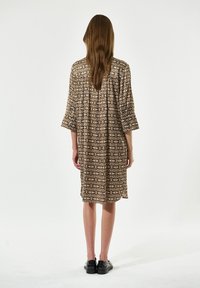 Braun gemustertes Kleid mit drei Viertelärmeln und lockerer Passform, das ein geometrisches Design aufweist. Schwarze Schuhe runden das Outfit ab.