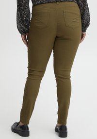 Olivnozelene skinny hlače iz raztegljive tkanine, z gladko teksturo, zadnjimi žepi in srednjim pasom. Oblečene z črnimi čevlji.