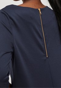 Haut bleu marine avec un col rond et une fermeture éclair dorée verticale à l'arrière, doté d'un tissu lisse et d'une coupe ajustée.