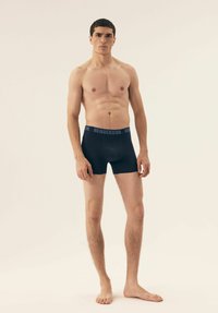 Black boxer shorts with a blue "HENDERSON" waistband, made from stretchy material, featuring a fitted design and smooth texture.