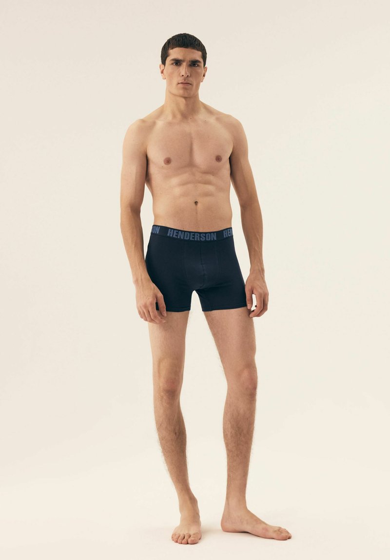 Black boxer shorts with a blue "HENDERSON" waistband, made from stretchy material, featuring a fitted design and smooth texture.