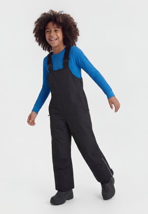 Bambino sorridente con capelli ricci che indossa una maglietta blu a maniche lunghe, salopette imbottita nera e stivali invernali neri, in piedi su uno sfondo bianco.