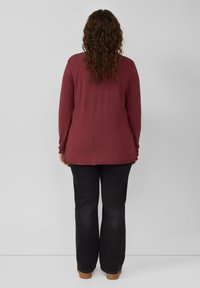 Langärmliges dunkelrotes Oberteil mit lockerer Passform, abgerundetem Saum und strukturiertem Stoff, kombiniert mit schwarzer, weit geschnittener Jeans und braunem Schuhwerk.