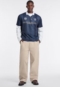 Navy short-sleeve polo shirt with white collar and stripes, featuring a logo and "GUESS JEANS" text. Paired with cream-colored long pants.