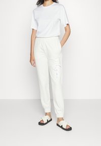 Bílá tričko spárované s světle barevnými tepláky s multikolorovým písmem na noze, doplněné černými šlapkami.