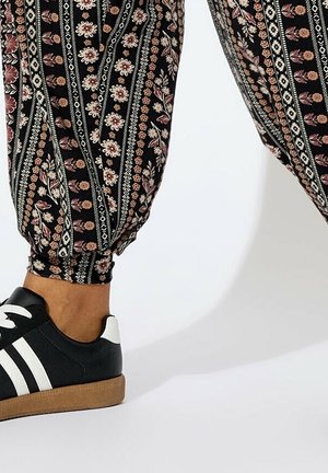 Benen met een zwarte en roze gebloemde broek met elastische boorden en zwarte sneakers met witte strepen en rubberen zolen op een witte achtergrond.