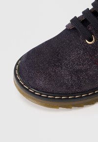 Donkerpaars suede schoen met een glinsterende textuur, ronde neus, zwarte veters, gouden oogjes en een contrasterende lichtgekleurde zool met stiksel detail.