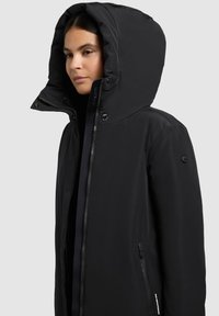 Naine, kes kannab musta kapuutsiga talvejope, millel on lukuga ja nööbiga üksteisega seotud detailid, on pööratud vasakule, helehallil ühevärvilisel taustal.