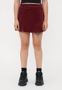 Bordeaux corduroy mini-jurk met een belted taille, voorzien van een split aan de voorkant en zichtbare stiksels. Draagt met zwarte schoenen.