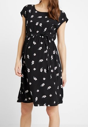 Robe de maternité noire longueur genou avec motif floral blanc et ceinture en tissu à la taille, portée par une femme enceinte.