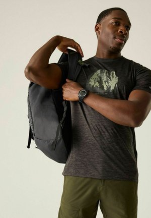 Man wearing a graphic t-shirt and watch puts on a black backpack over his shoulder against a plain background.