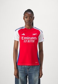 Červeno-bílý sportovní dres s krátkým rukávem a modrými akcenty, se znakem "Emirates Fly Better", kulatým výstřihem a třemi pruhy na ramenou.