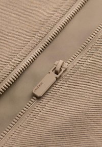 Beżowa tkanina z zamkiem błyskawicznym, charakteryzująca się teksturowanym wzorem w paski. Uchwyt zamka jest w stonowanym odcieniu, wytłaczanym z nazwą marki "ambre."