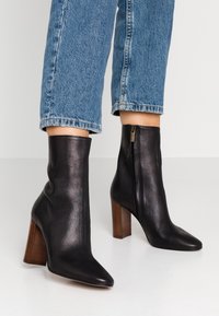 Black leather ankle boots with a pointed toe, high block heel, and side zipper. Smooth texture with a structured design.