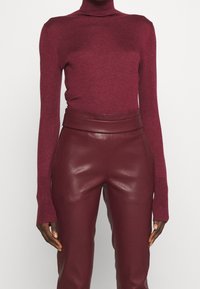 Suéter burdeos de cuello estilo chimenea, hecho de un suave tejido de punto con mangas largas, combinado con pantalones de piel burdeos de cintura alta que tienen un acabado liso.