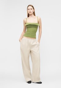Γυναίκα που στέκεται με τα χέρια στις τσέπες, φοράει πράσινο αμάνικο τοπ και φαρδιά μπεζ παντελόνια, μπροστά σε απλό φόντο.