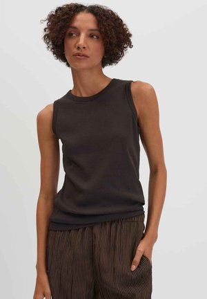 Donna con capelli ricci che indossa una canotta nera e pantaloni marroni a righe, in piedi con una mano in tasca, guarda verso sinistra.