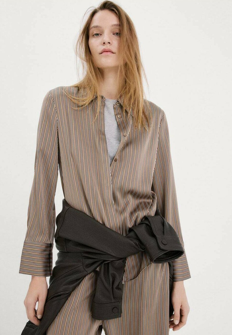 Mlada žena s dugom plavom kosom, odjevena u smeđu prugastu košulju na kopčanje, sivu unutarnju majicu i crnu jaknu vezanu oko struka.