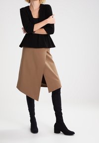 Zwarte peplum top, tan asymmetrische rok met een zijslit, en kniehoge zwarte laarzen. Stof lijkt glad en gestructureerd.