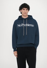 Námořnická modrá mikina s kapucí a bílým textem "No Problemo", s tkaničkami v kapuci a žebrovanými manžetami, nošena spolu s černými kalhotami. Hladká textura.