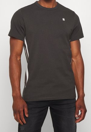 Homme portant un t-shirt gris foncé à manches courtes avec des bandes blanches sur les côtés et un jean noir, debout devant un fond uni.