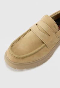 Pantof cu slip-on din suede bej, cu vârful întărit și cusături decorative, având o talpă texturată pentru aderență și un accent de marcă pe bandă.