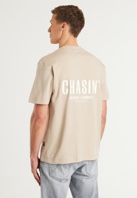 Beige bomulds t-shirt med korte ærmer og rund halsudskæring, med hvid tekst på ryggen: "CHASIN' A1S BASED C.COMMUNITY." Enkel design.