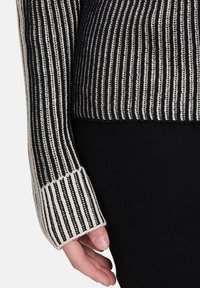 Ręka w czarno-biały pionowo paskowany sweterdługi rękaw, odpoczywająca obok czarnych spodni na jednolitym tle.