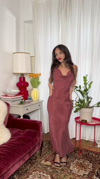 Γυναίκα με μωβ αμάνικο φόρεμα και μαύρες γόβες στέκεται σε σαλόνι με κόκκινο βελούδινο καναπέ, τραπεζάκια, φυτά και λευκές κουρτίνες.