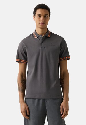 Young man wearing a gray polo shirt with orange striped collar and sleeve cuffs, standing against a plain white background.