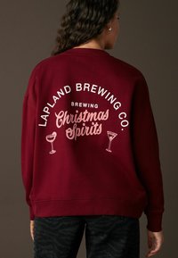 Karmīnsarkans džemperis ar brīvu piegriezumu, ar baltu un rozā tekstu, kurā teikts "Lapland Brewing Co. Ražojot Ziemassvētku Garus", kā arī kokteiļu grafika.