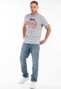 Grå bomulds t-shirt med en rød og blå grafisk tryk med en løve og tekst. Sammen med blå denim jeans og hvide sneakers.