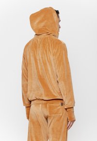 Bluza z kapturem z miękkiego, jasnobrązowego materiału. Charakteryzująca się luźnym krojem, ściągaczami przy mankietach oraz detalem kieszeni na plecach. Gładka tekstura i prosty design.