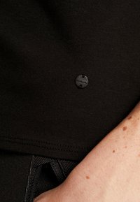 Sort bomulds T-shirt med et lille rundt logo mærke. Stoffet er glat med en let glans, og syningerne er pænt afsluttet.