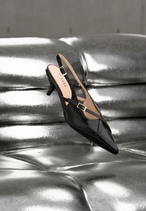 Czarna lakierowana skórzana czółenka na niskim słupku typu slingback z złotą klamrą, spoczywająca na błyszczącej srebrnej pikowanej tapicerce skórzanej.