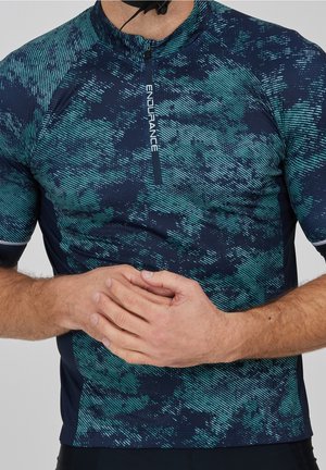 Mann trägt ein blau-grün gemustertes kurzärmliges Sportoberteil mit "ENDURANCE" am Reißverschluss, Hände vor der Taille verschränkt.
